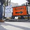 A low-angled photo shows peoples legs as they stand on a sidewalk by a large orange sign reading "testing"