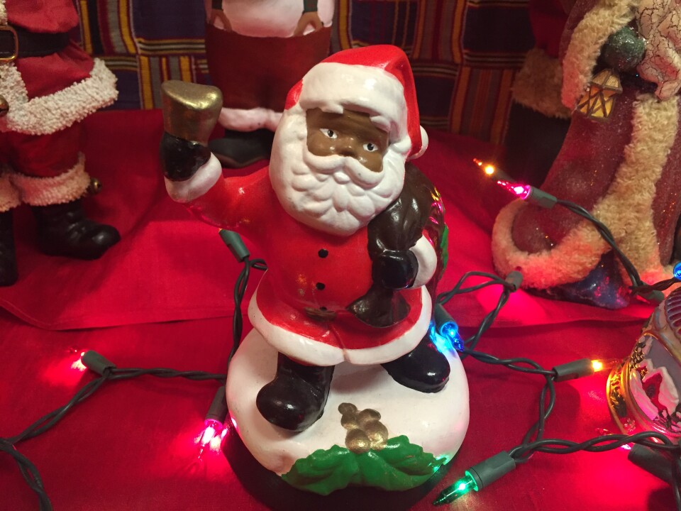 A small figurine of a Black Santa Claus sits on a table surrounded by multicolored string lights and other figurines.