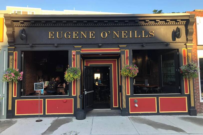 This is an exterior shot of the Eugene O'Neill's pub. It's a sunny day. It must be warm as the front door to the pub is propped open. The exterior is painted black and red. Four flowering hanging baskets decorate the outside of the pub. A few people are seated at the bar, reading a menu.