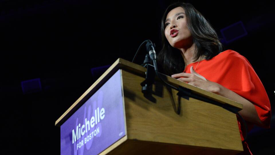Boston Mayor Michelle Wu is wearing a red blouse, standing at the podium and addressing a crowd.