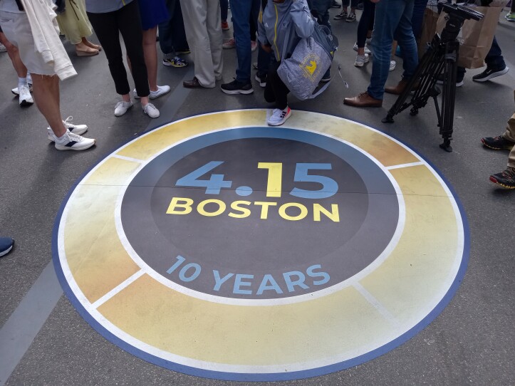 A circular, blue, yellow and white marker with the words "4.15 Boston, 10 Years" sits on the ground near the Boston Marathon finish line. The number one in the 4.15 and Boston are highlighted to read "One Boston"
