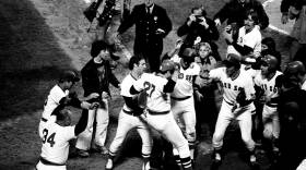 A baseball player jumps with both feet onto home plate as a crowd of teammates cheer around him.