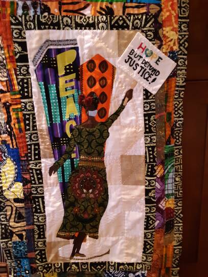 A quilt with bright patterns shows a Black woman raising a sign that reads "Hope but demand justice!"
