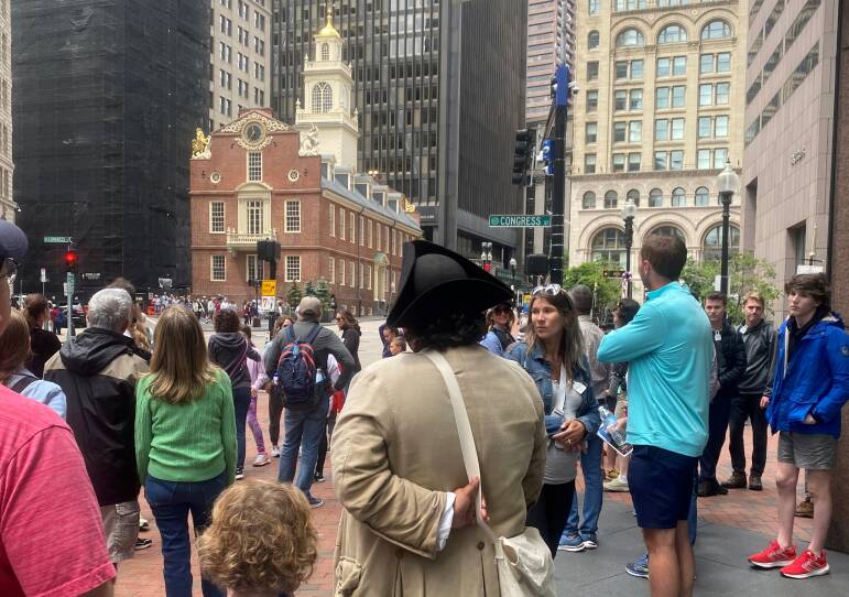 A man in a colonial costume including a tri-cornered hat stands with a group of tourists in front of Boston's Old State House as he describes the history of the building on the Freedom Trail.