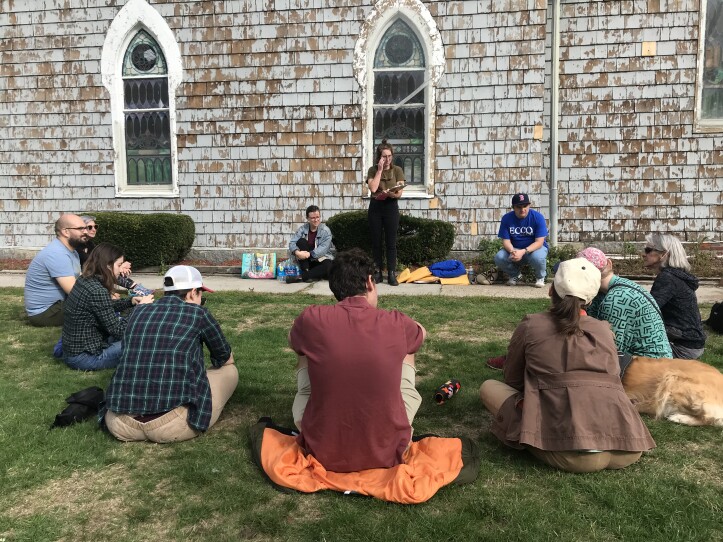 A young woman stands while a small group of 10 to 12 people sit in a semicircle around her, taking instructions. They’re sitting on a lawn in front of a church with peeling paint.