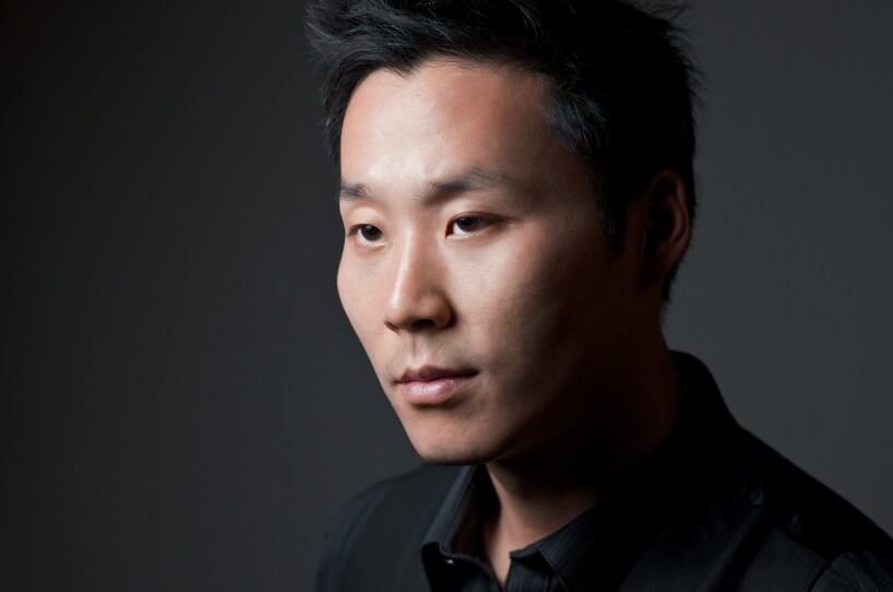 This is a headshot of a young man. He has short black hair, he's wearing a black, opened collar shirt. He is facing left.