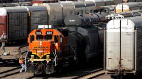 A rail yard with many lines and freight cars stretching out into the distance. In the foreground, a train engine, painted orange with bold black and yellow stripes, appears as if it's being pulled by a rail worker wearing a ballcap, jeans, and a safety vest.