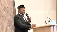 A Black man wearing a suit, bow tie and a religious cap of the Nation of Islam stands at a podium and speaks with a hand-held microphone.