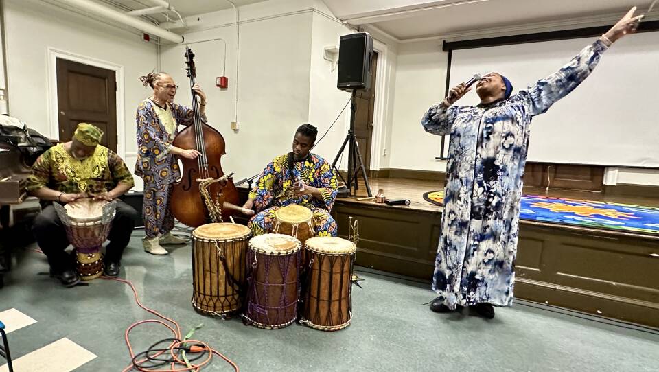 A Black woman in a long blue and white printed dress sings into a microphone with one arm pointing in the air while 3 musicians play drums and bass nearby.