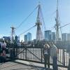 Two people stand behind a metal fence as they look at an old wood-masted sailing ship.