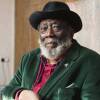 A portrait of artist Frank Bowling, wearing a green suit jacket, red button up shirt, and black hat. Bowling looks at the camera and sits in front of a beige wall.