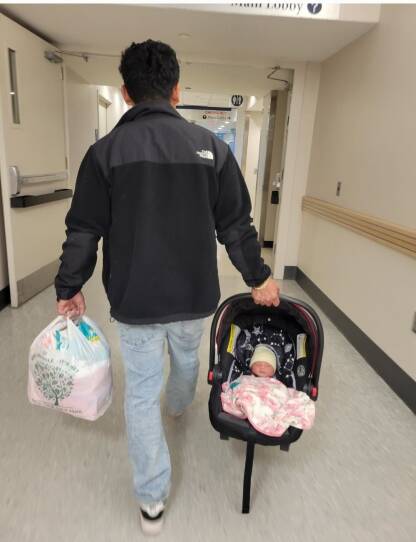 A man walks down a hospital corridor holding an baby car seat with an infant in it.