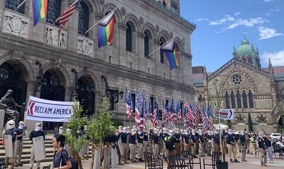 A group of men in khaki pants and face masks hold up American flags and a sign that says "Reclaim America." They stand in front of the Boston Public Library.
