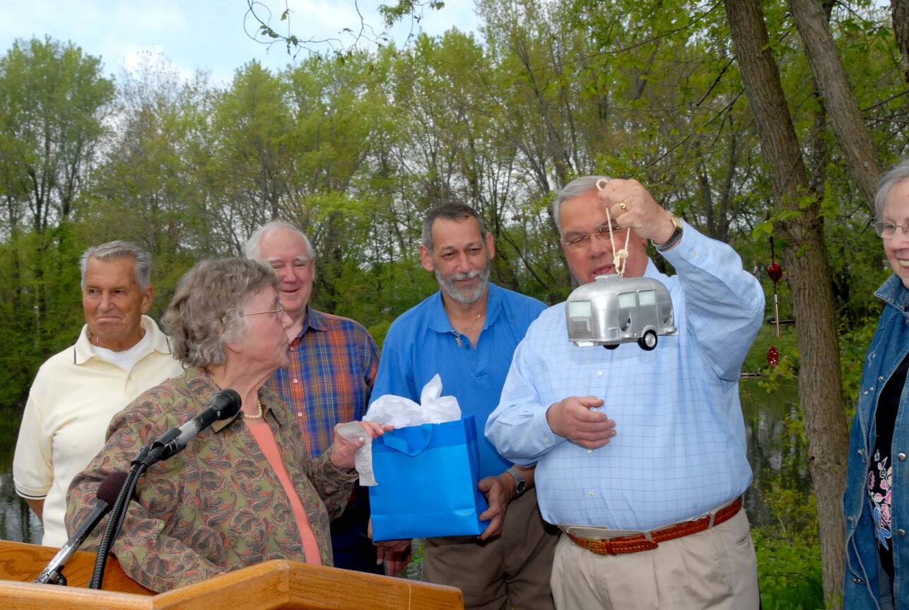 A man in a checkered blue shirt excitedly holds up a miniature gray trailer home while others look on, one holding a gift bag