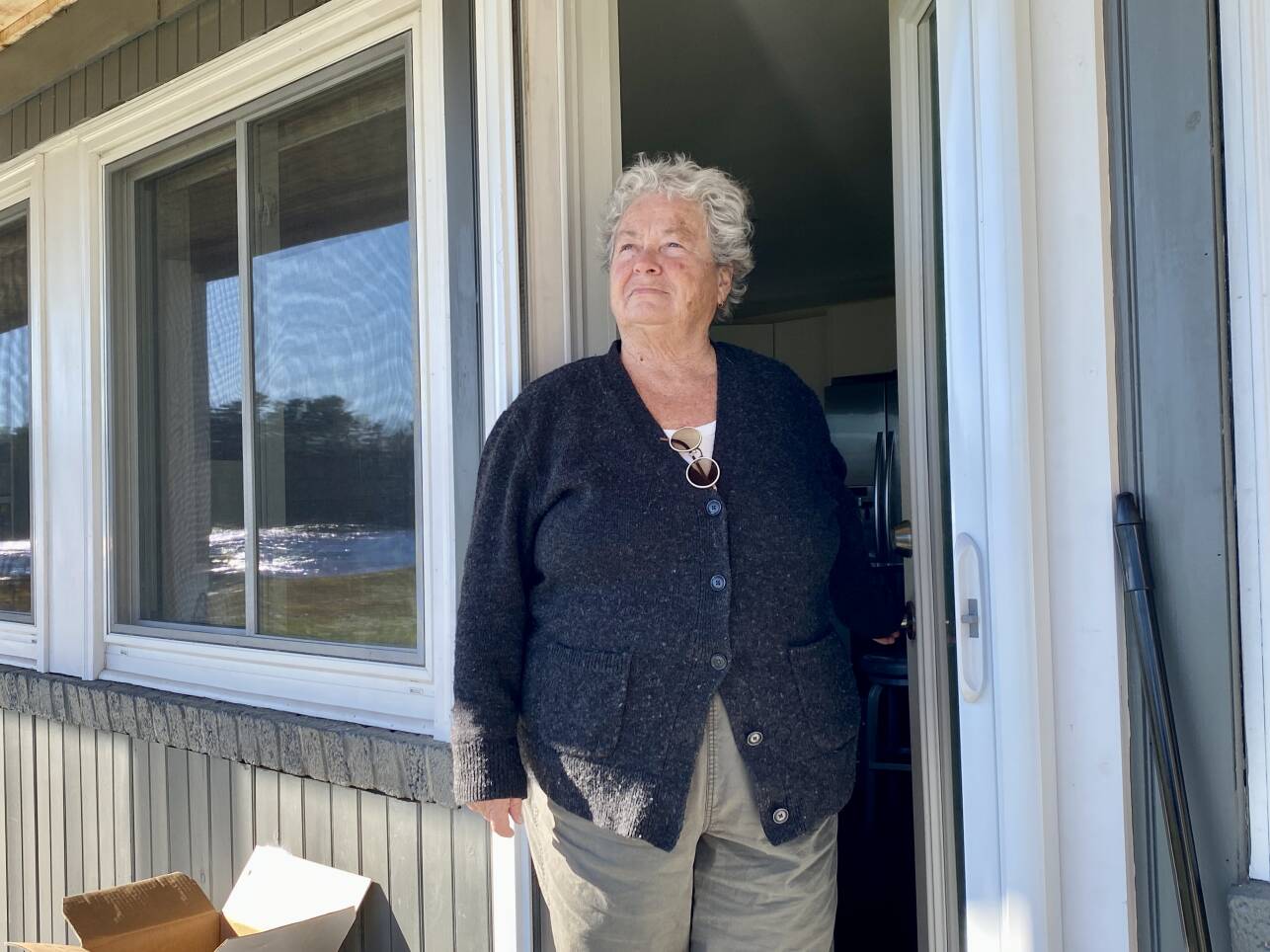 A white woman with short silver hair stands in the doorway of a home and looks out at the horizon with a river reflected in the window nearby