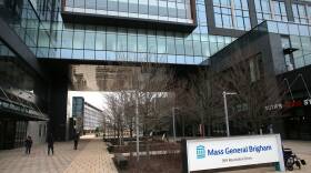 A few people walk under a pedestrian tunnel in a large glass building, a sign reading "Mass General Brigham" out front