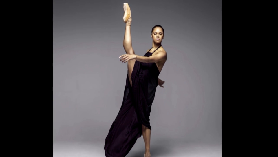 A ballerina is dressed in a sleeveless black dress. She is kicking her right leg up vertically so that it extends beyond her head. She extends her left arm outward,  her right arm is extended behind her back.