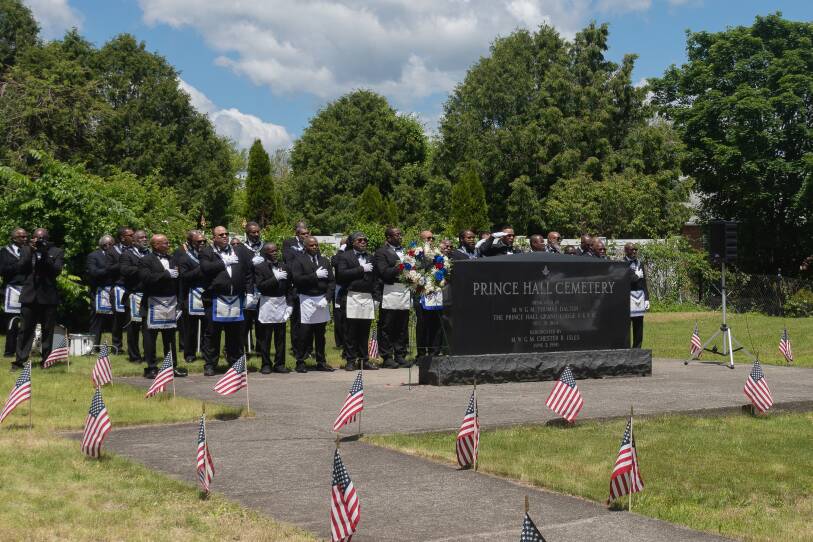 A group of Prince Hall Freemasons in their Masonic attire wearing Black tuxedos and white aprons holding their hands on their chest during the Pledge of Alliance. They are standing behind a large black tombstone that reads "Prince Hall Cemetery."