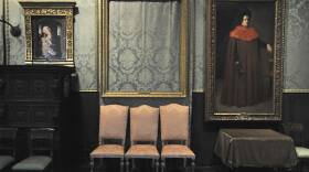Three chairs sit underneath an empty golden frame that once held a painting, flanked by two other paintings in gold frames.
