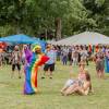 A drag performer Natasha Star wearing a bright rainbow gown and multicolor wig sings during a festival in a grassy town park. Dozens of people in the park watch, clap and hold up their phones to take photos.