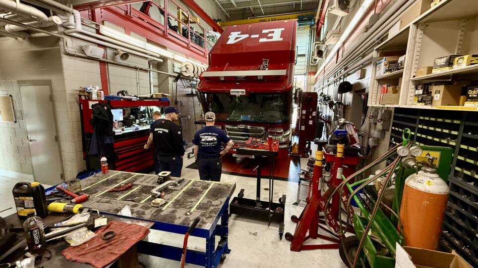 A wide shot of a repair shop with a Woburn fire truck being serviced. Two mechanics are in foreground talking about the repairs.
