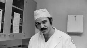 A black and white photograph of a man wearing a doctor's uniform in a hospital exam room.