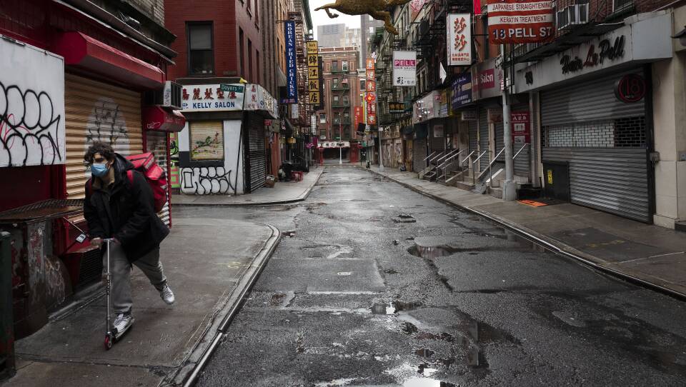 A person on a scooter moves along the sidewalk of an empty street surrounded by restaurants with boarded up windows.