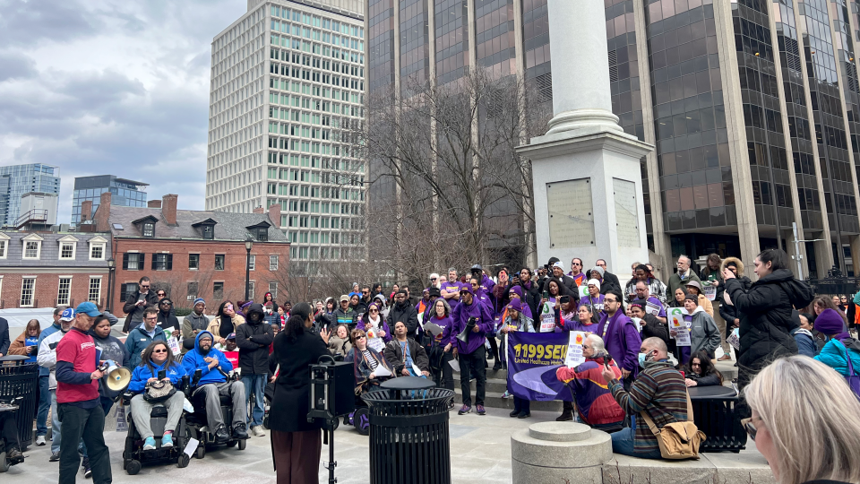 Dozens of people gather, some in wheelchairs, listening to a person at a podium.
