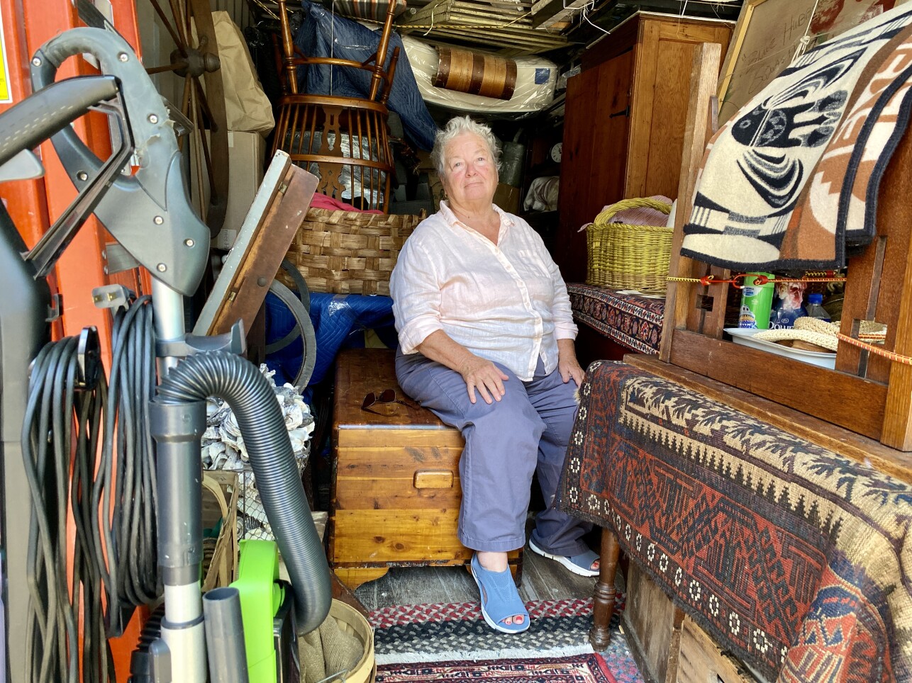 An older woman with short white hair sits on a wooden chest surrounded by stacks of her belongings.