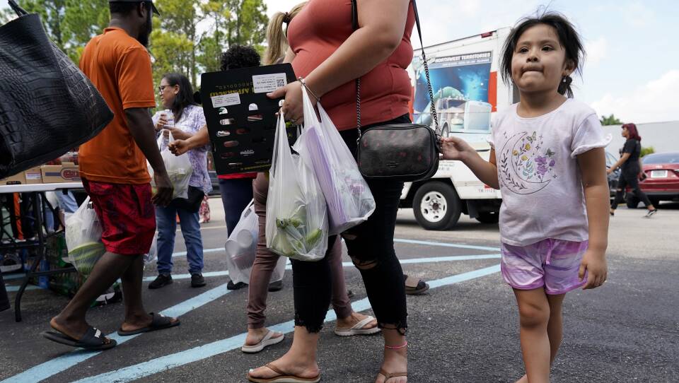 A young girl holds onto the strap of her mother's purse as the mom carries a box and two plastic bags.