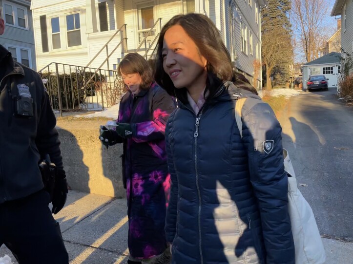 A woman in a winter coat walks on the sidewalk outside of a house. A police officer and another woman are beside her.