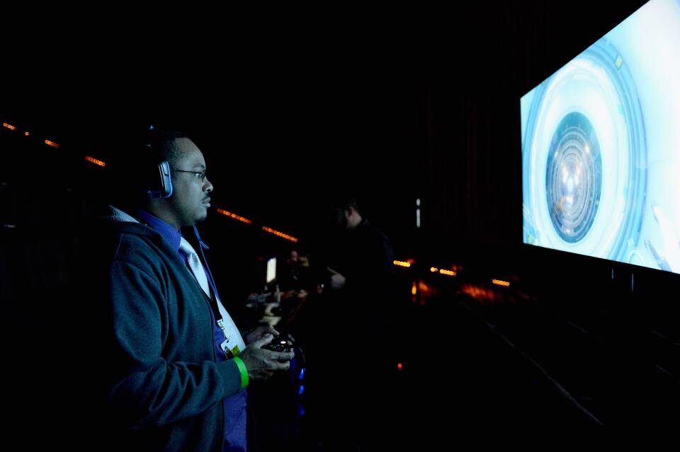 A man with large headphones stands in a dark room, playing a video game.