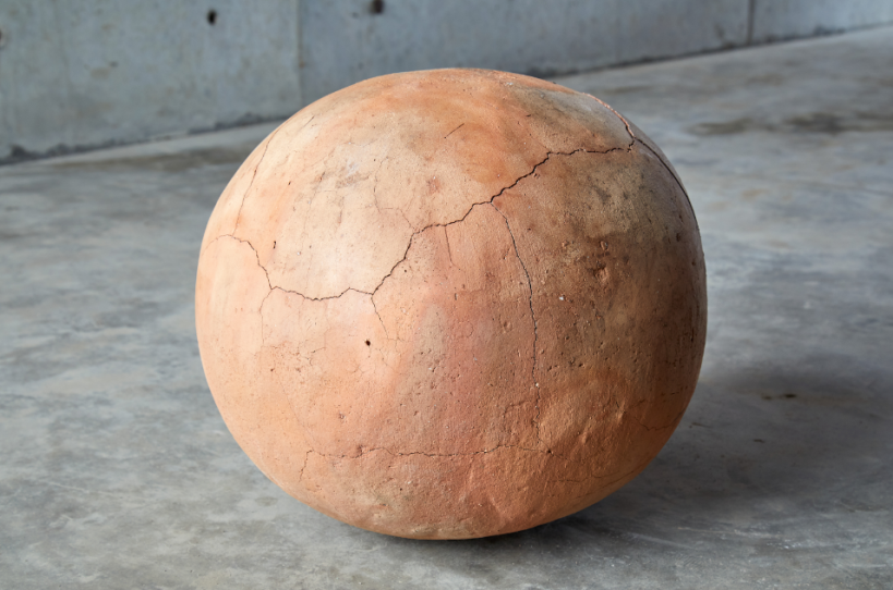 A clay sphere with a large crack running down is sits on a concrete floor