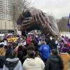 A crowd of people stand and cheer in front of the Embrace statue. They hold flags and signs that read '1199 SEIU.'