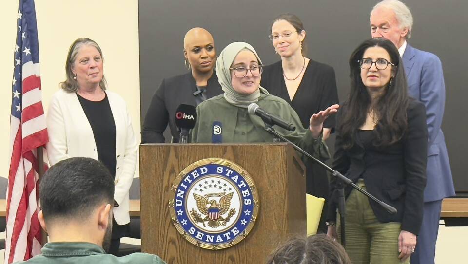 A woman stands at a podium, speaking into a microphone. Behind her are five people in business attire.