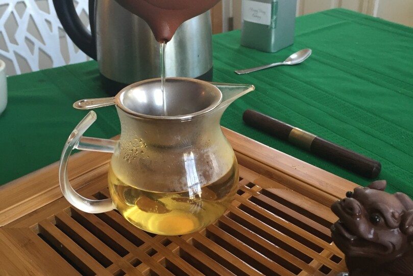 Oolong tea being strained into a glass carafe, on a wooden tea tray .
