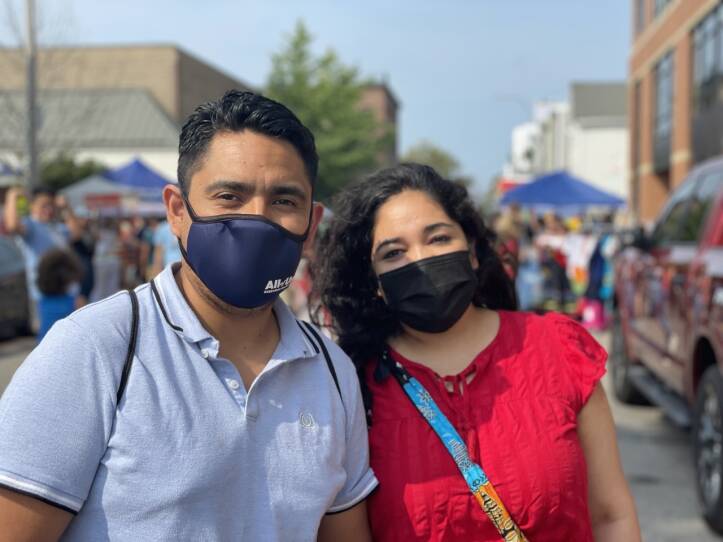 A man and woman wearing masks stand and pose on a busy East Boston street