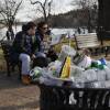 A trash can on the edge of a paved walkway overflows with plastic cups, bottles and other trash. A couple sits on a bench behind it.