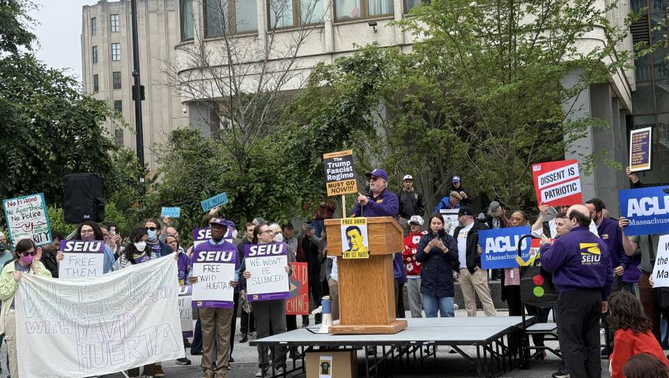 A man speaks behind a podium and other people around him hold up signs with political messages.