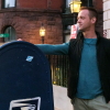A man in a sweater and light fleece vest stands leaning against a mailbox on the sidewalk of a Boston neighborhood.