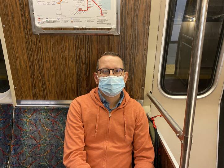 A man in a blue surgical mask, glasses, and an orange zip-up hoodie sits on the Orange Line.