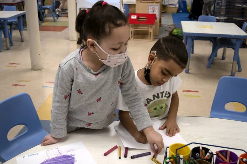 Two young children draw with crayons while standing at the edge of a table in a daycare classroom.