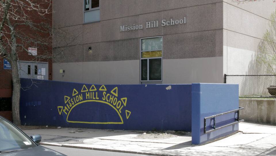 A bright blue wall has "Mission Hill School" and the outline of the sun painted on it in bright yellow, in front of a ramp to the school's entrance