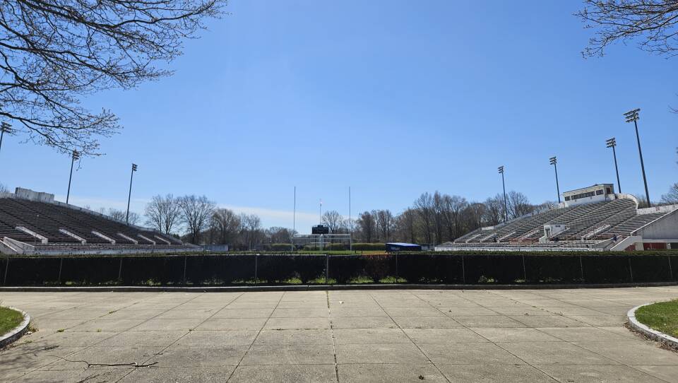 A stadium with rows of bleachers and stadium lights is shown between two trees and underneath an open, blue sky.