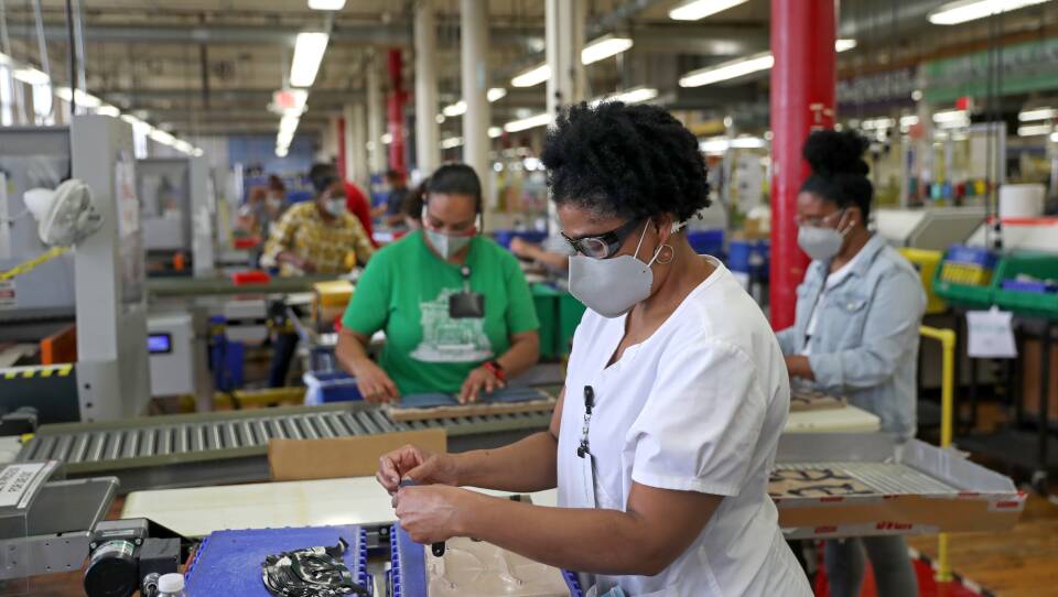 A woman stands in a factory in the foreground, wearing a T-shirt and a protective mask. She’s making something small. Other women can be seen in the background doing the same task.