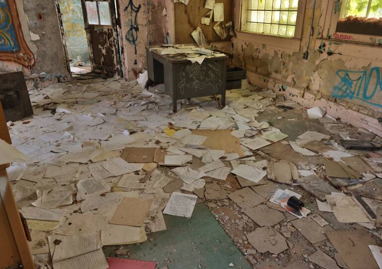 Old documents and papers are strew across the floor of a run-down building. Graffiti is on the walls.