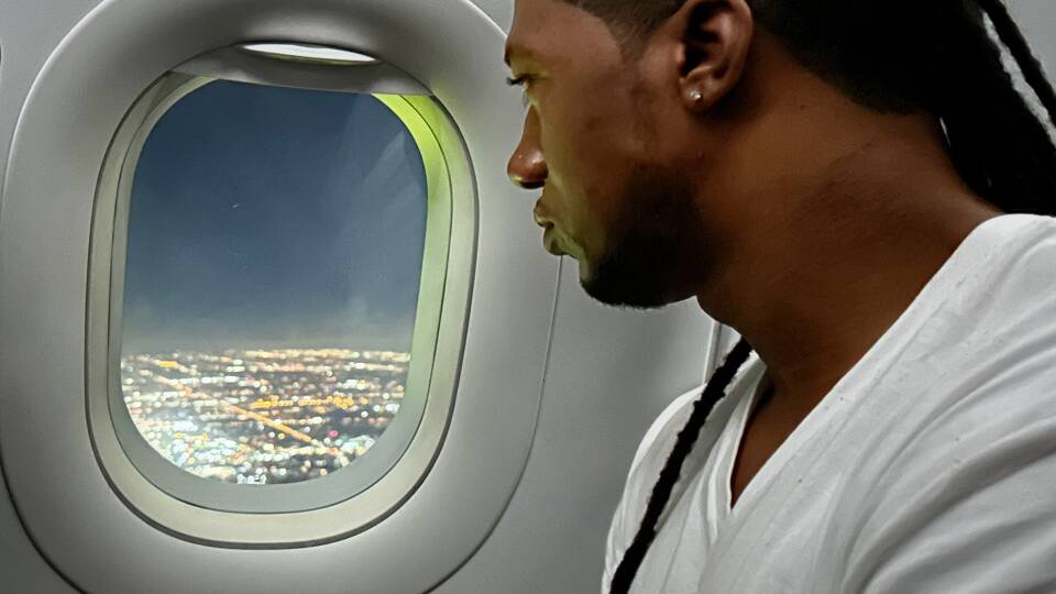 A man with a few hanging dreadlocks looks out the window of a plane.