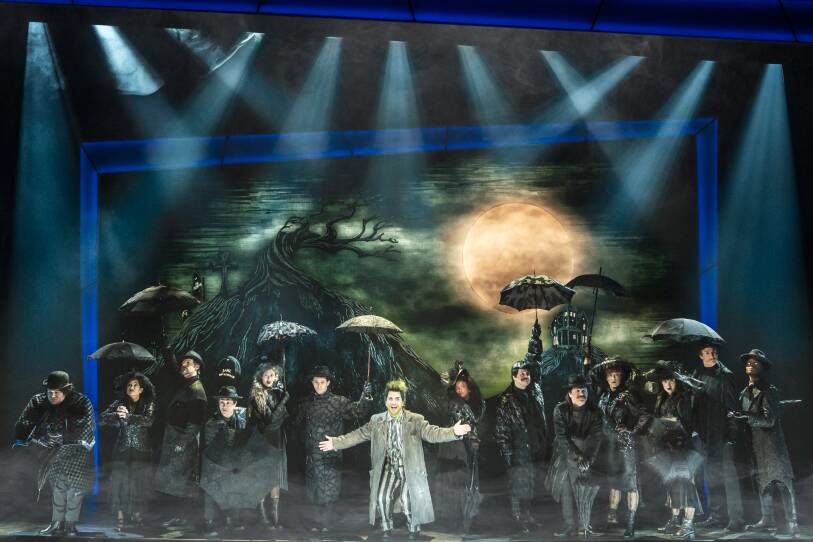 the cast of "Beetlejuice" perform at the Citizens Bank Opera House. Everyone onstage, except for the titular character in the center, wears all black and holds black umbrellas. They stand in front of a dark green backdrop depicting a nighttime scene