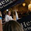 A woman in a white pantsuit beams out over a crowd, viewed between signs supporting her candidacy. She stands in front of Massachusetts state flags in an ornate room with a large chandelier hanging.