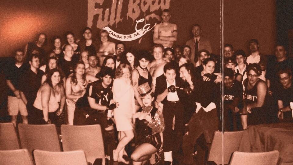A black and white photo of a group of performers standing at the front of a theater, dressed in costumes from "The Rocky Horror Picture Show."
